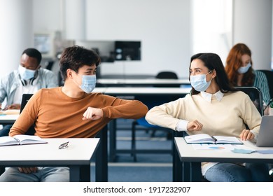 Elbow Bump. Young Multiethnic Students Wearing Protective Disposable Surgical Masks Greeting Each Other At Classroom. Man And Woman Avoid Touching, Studying In High School, Sitting At Table