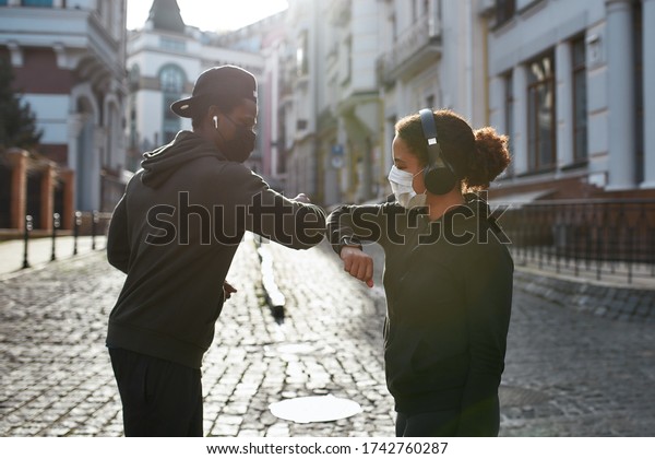 Elbow bumping. Young african couple of runners\
in medical protective masks bumping elbows instead of greeting,\
standing at the empty city street in the morning. Social distance.\
Coronavirus epidemic