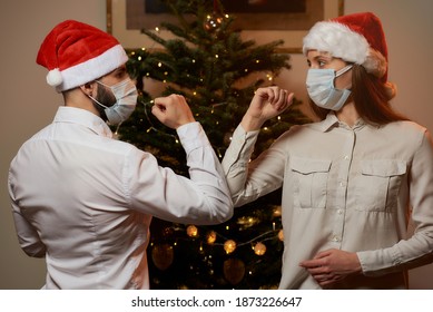 Elbow Greeting To Avoid The Spread Of Coronavirus (COVID-19) Against A Background Of Christmas Tree At Home. Man And Woman In Medical Face Masks And Santa Hats Instead Of Hug Or Handshake Bump Elbows