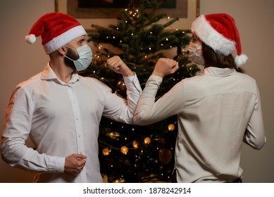 Elbow Greeting To Avoid The Spread Of Coronavirus Against The Background Of A Christmas Tree At Home. A Man And A Woman In Medical Masks And Santa Hats. Instead Of A Hug Or Handshake, They Bump Elbows