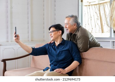 Elder Asian Man And His Son Using Video Call From Mobile Phone To Say Hi To The Rest Of Family In His Cozy Retirement Home