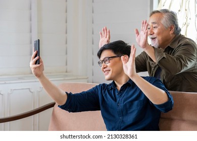 Elder Asian Man And His Son Using Video Call From Mobile Phone To Say Hi To The Rest Of His Family In Cozy Retirement Home