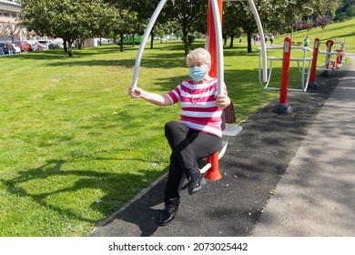 Elder Blond Woman With Face Mask Doing Exercise On Park Machine On Sunny Day. Senior Lady Training Upper Body To Keep Fit And Healthy Outdoors