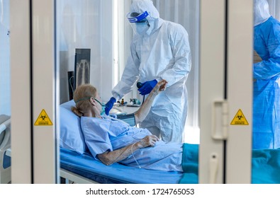 Elder Caucasian Female Patient Gets Blood Pressure Checked By Team Of Medical Doctors In Quarantine Room. Medical Staff Wear Sterile Personal Protective Equipment Suit And Face Shield. COVID-19 Check