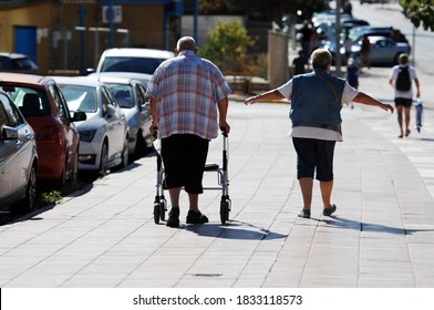 An Elder Couple Walking On A Street In Finestrat-Spain. An Overweight Man With A Walking Aid. An Older Woman Spreads His Arms.