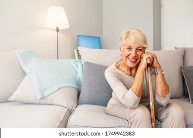 Elder lady sitting on the couch with wooden walking stick and smiling. Happy elderly woman relaxing on sofa and holding walking stick. Shot of a senior woman looking thoughtful in a retirement home