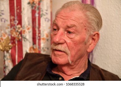 An Elder Man Withblue Eyes And A Walrus Mustache Is Listening To A Conversation.