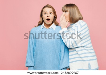 Elder parent mom 50s year old with young adult daughter two women together wear blue casual clothes whisper gossip and tells secret behind her hand isolated on plain pink background Family day concept