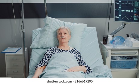 Elder Patient With IV Drip Bag And Nasal Oxygen Tube Laying In Bed. Aged Woman With Disease Receiving Healthcare Treatment For Recovery In Hospital Ward, Waiting For Medical Support.