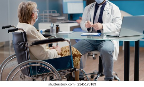 Elder Patient With Physical Disability Talking To Doctor At Checkup Appointment In Cabinet. Senior Woman In Wheelchair Receiving Medical Treatment To Help With Rehabilitation.