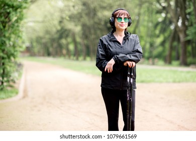 Elder Woman In Black Sportswear Walking With Hiking Sticks In The Park