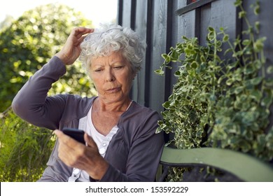 Elder Woman Sitting On A Bench In Her Backyard Reading Text Message On Her Mobile Phone