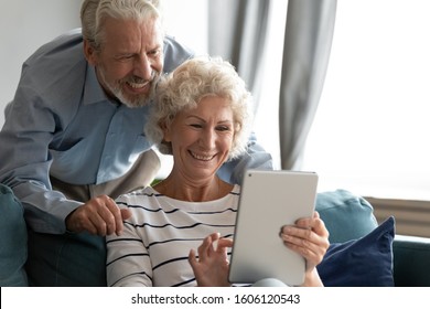 Elderly 60s Couple Spend Time At Home, Wife Holding Tablet Buy Or Ordering Products Through Virtual Space Via Internet, Easy Quick Services, Older Generation Active Users Of Up To Date Devices Concept