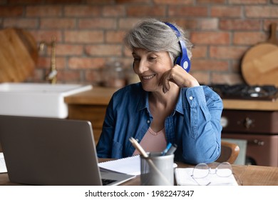 Elderly 60s grey-haired woman in headphone sit in home kitchen participate in video call conversation use computer application. Older citizen and modern technology usage, distant communication concept