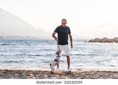 Elderly 60-years Old Man Playing With Small Cute Jumping Funny Dog Jack Russell Terrier On Beach