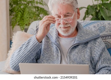 Elderly 70s Man Seated On Sofa Browsing On Laptop, Senior Looking At Laptop Screen Surfing The Net, Older Generation And Modern Tech Application Easy Usage Concept