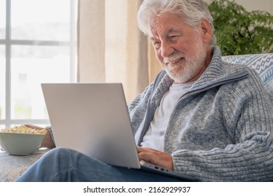 Elderly 70s Man Seated On Sofa Browsing On Laptop, Senior Looking At Laptop Screen Surfing The Net, Older Generation And Modern Tech Application Easy Usage Concept