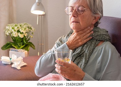 Elderly Adult Caucasian Woman With Sore Throat And Fever As Seasonal Flu Or Pollen Allergy On Sofa At Home While Drinking Glass Of Water