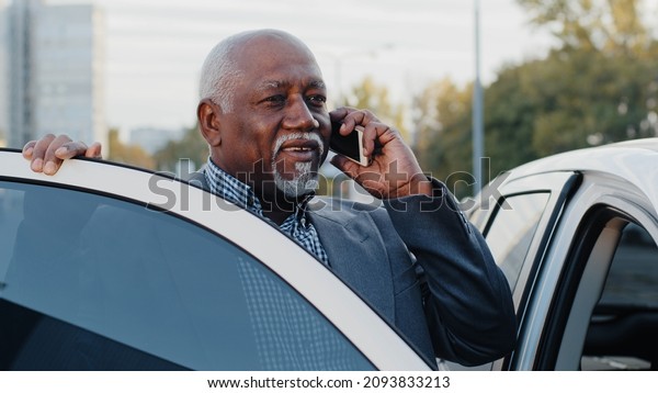Elderly african american man talking on mobile\
phone walks up to car sits down in front passenger seat answers\
call using smartphone mature businessman discussing business\
conversation on\
telephone