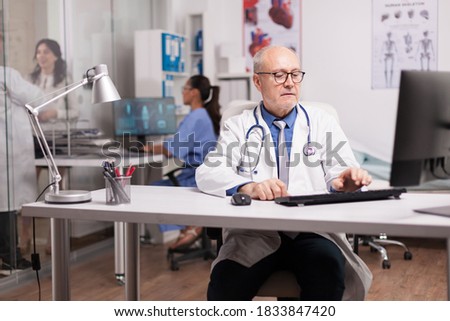 Elderly aged doctor using computer in hospital cabinet while young doctor talking with female patient on clinic corridor and nurse typing rapport on pc.