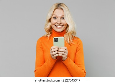 Elderly amazed shocked satisfied fun happy blonde caucasian woman 50s in orange turtleneck hold in hand use mobile cell phone isolated on plain grey color background studio People lifestyle concept
