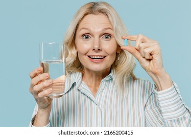 Elderly Amazed Woman 50s Wear Casual Striped Shirt Hold Glass Of Water Medication Tablets, Aspirin Pills Isolated On Plain Pastel Light Blue Color Background Studio Portrait. People Lifestyle Concept