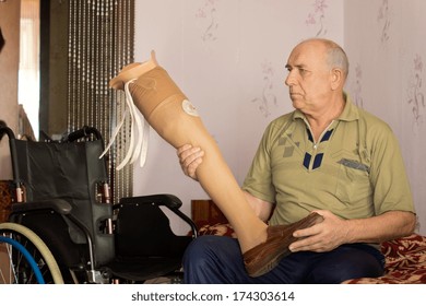 Elderly Amputee Sitting On His Bed Alongside His Wheelchair Holding An Artificial Leg In His Hand As He Prepares To Fit It To His Stump