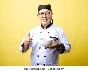 An elderly Asian chef in a white uniform and black hat smiles while holding a bowl and giving a thumbs-up against a bright yellow background - Powered by Shutterstock