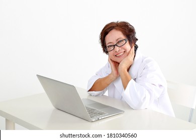 Elderly Asian Chinese Female Doctor On White Background Wearing White Apron Notebook Computer Hand On Cheek Happy Look At Camera