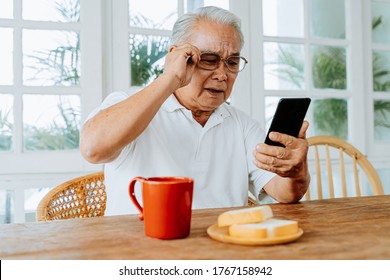 Elderly Asian Male Having Eyesight Problems And Wearing Eyeglasses While Gazing And Reading Message On Smartphone During Breakfast At Home. Old Man Having Eye Blurred Vision Indoors.