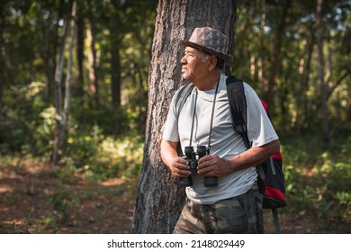 An Elderly Asian Man With Backpack Looking At Binoculars In The Forest.