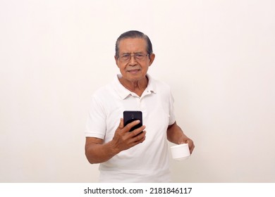 Elderly Asian Man Standing While Holding His Cell Phone And Cup. Isolated On White Background