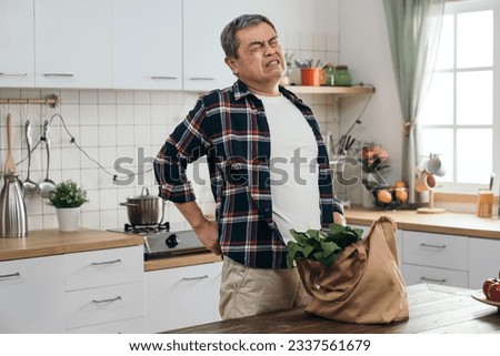 elderly Asian man strains his back carrying a heavy shopping bag with healthy food ingredients from the market or store, mindful of his health.