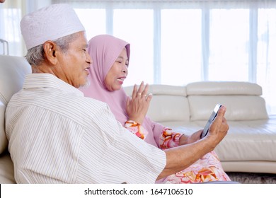 Elderly Asian Muslim Couple Holding A Phone, While Talking To Their Grandchildren Via Video Call In Their Living Room