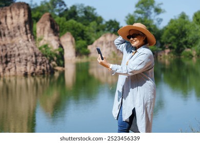 Elderly Asian woman adjusting hat while taking a selfie near a tranquil lake, surrounded by nature and cliffs. The sunlight enhances the calm and relaxed mood of the outdoor setting. - Powered by Shutterstock