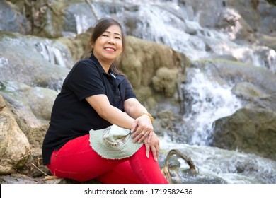 Elderly Asian Woman In A Black Shirt Is Touring The Natural Beauty Of The National Park, Thailand. Asian Woman Are Sitting On The Floor With Blurred Of Waterfall Background.