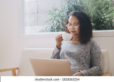 Elderly Asian Woman Drinking Coffee And Working From Laptop At Her Room.