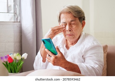 Elderly Asian woman with grey hair is using mobile phone and rubbing her eyes with her hand. Eye problems in the elderly, blurry vision, concept