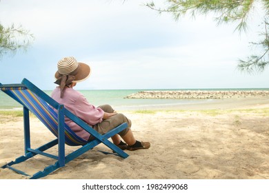 An elderly Asian woman in a hat sits on a deck chair by the beach. The concept of the elderly travel nature during retirement age. Copy Space
