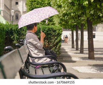 An Elderly Asian Woman Sits On A Bench Under The Sun With A Purple Umbrella