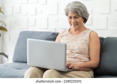 Elderly Asian Woman Typing Laptop Computer While Sitting On Sofa At Home