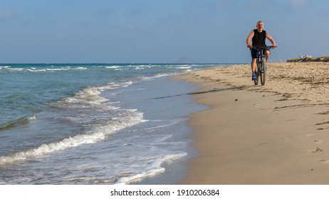 An elderly athlete rides his mountain bike across the beach near Torrevieja. The waves from the Mediterranean roll onto the sand. It is just before sunset in the afternoon.