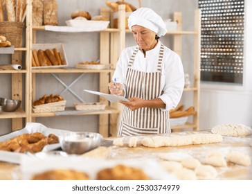 Elderly baker woman standing at a bakery table, holding a piece of paper and pen, planning and organizing tasks. - Powered by Shutterstock