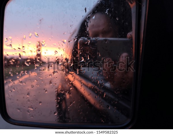 An elderly bald man\
photographs himself on smartphone through mirror of car covered in\
raindrops at sunset