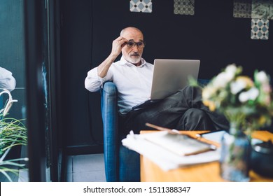 Elderly Bald Man In White Shirt And Black Pants Correcting Glasses And Working On Laptop While Sitting In Living Room