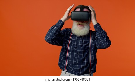 Elderly bearded gray  haired man using virtual reality futuristic technology VR app headset helmet to play simulation 3d 360 video game  drawing  Orange studio background  Senior grandfather pensioner