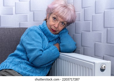 An Elderly Beautiful Woman Of 60-65 Is Warming Up At The Heating Radiator In Her Room, Looking At The Camera. Concept: Low Temperature In The Room, Rising Gas And Heating Prices, Autumn Cold Snap.