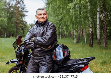 Ciclista de ancianos apoyándose en motobike en el bosque de verano. Hombre con ropa de cuero parado con los brazos cruzados disfrutando la naturaleza. después del viaje