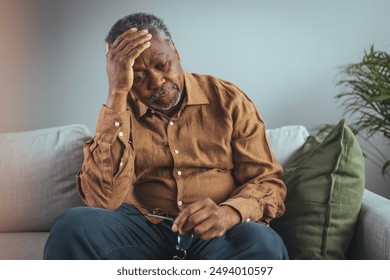 Elderly Black Man Experiencing Discomfort on Couch. Senior black male grips his glasses, showing signs of distress while seated alone on a sofa, reflecting potential health concerns in a home setting. - Powered by Shutterstock