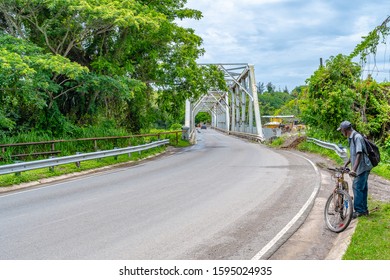 Elderly Black Man Standing With Bicycle On Street Entrance To Short Narrow Steel Bridge In Lush Rural Countryside Landscape On Caribbean Island. Wag Water Bridge In Junction, Saint Mary, Jamaica.